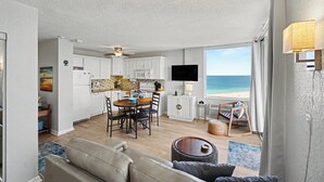 Eine moderne Küche mit weißen Schränken, ein Essbereich mit Tisch und Stühlen, ein Flachbildfernseher an der Wand und Blick auf den Strand.
