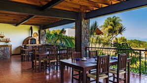 A covered outdoor dining area with wooden tables and chairs, a stone fireplace, and a view of greenery and mountains.