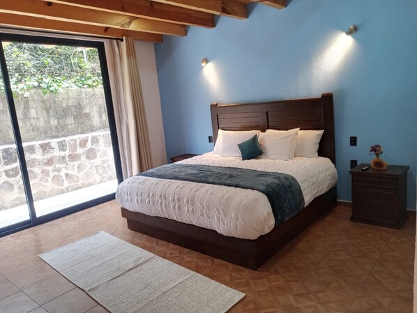 Un dormitorio con una cama grande, cabecera de madera, mesita de noche y vistas a un muro de piedra y follaje exterior.