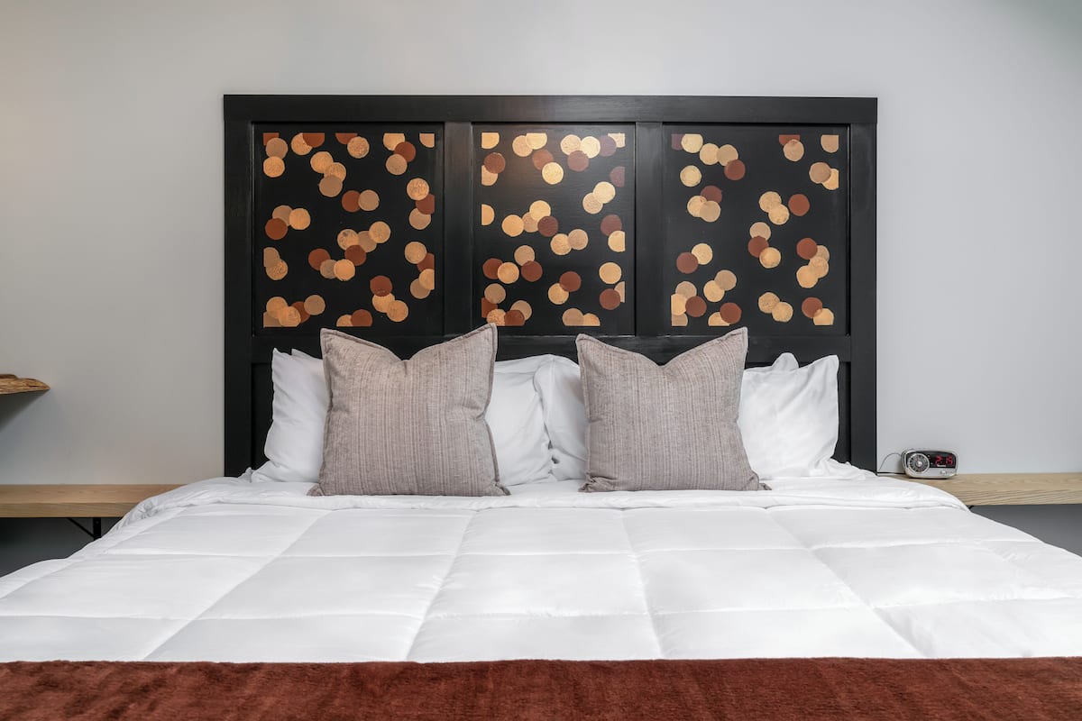 Ein ordentlich gemachtes Bett mit weißer Bettwäsche und zwei Kissen, ein Nachttisch aus Holz und eine an der Wand befestigte Uhr.