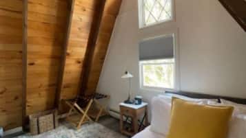 A cozy attic bedroom with a sloped ceiling, a bed with white and yellow bedding, a small table with a lamp, and a window with a view.