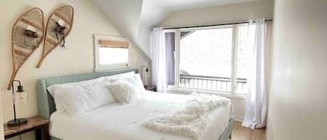 Une chambre à coucher avec un lit, un plancher en bois, une fenêtre avec des rideaux et un luminaire fixé au mur.