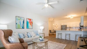 Una sala de estar moderna con un sillón de cuero marrón, una mesa de centro de vidrio y un sofá color crema. Se observa una cocina con armarios blancos y un refrigerador de acero inoxidable.