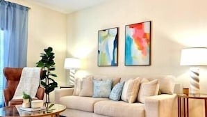 Un salón con un sofá beige, dos cuadros abstractos, una planta en maceta, una mesa de centro de vidrio y una lámpara.
