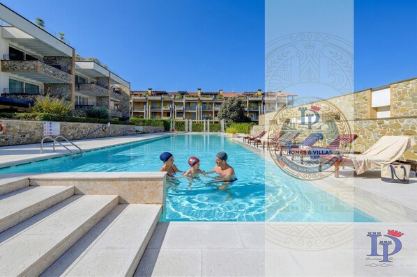 A swimming pool area with steps leading into the water, a stone wall, and a building with balconies in the background.