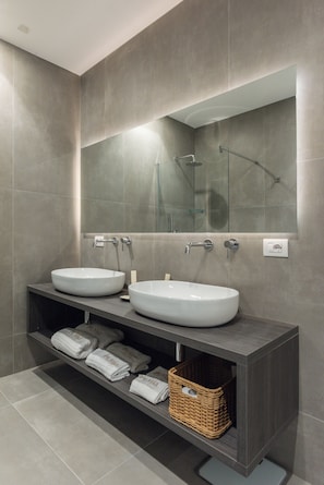 A modern bathroom with two sinks, a large mirror, and a shelf with towels and a basket.