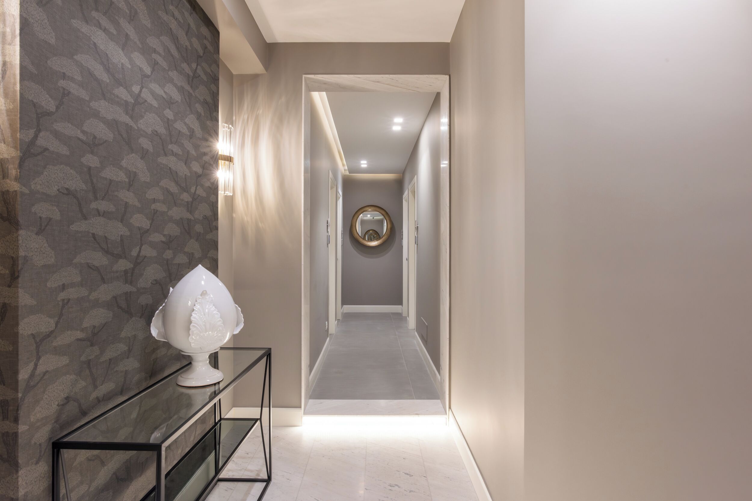 A hallway with a patterned wall, a white vase on a console table, and a round mirror on the wall.