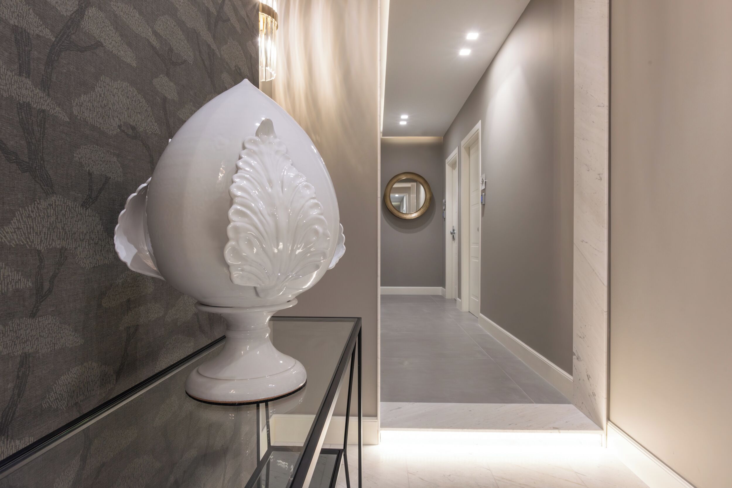 Um corredor com um objeto decorativo branco sobre uma mesa de vidro, um espelho redondo na parede e uma porta no final.