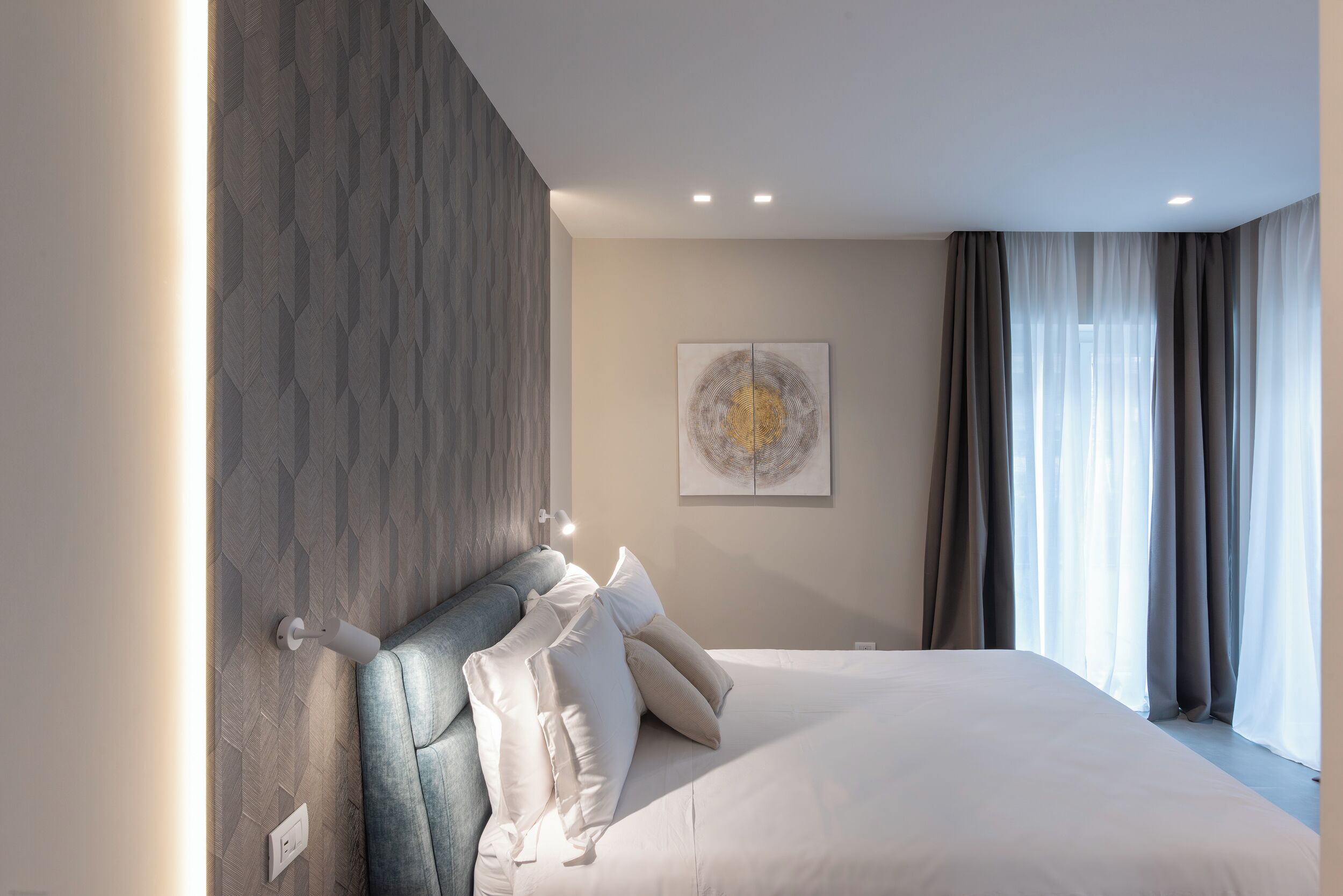 A modern bedroom with a large bed, a headboard with geometric patterns, a wall-mounted light, and a window with curtains.
