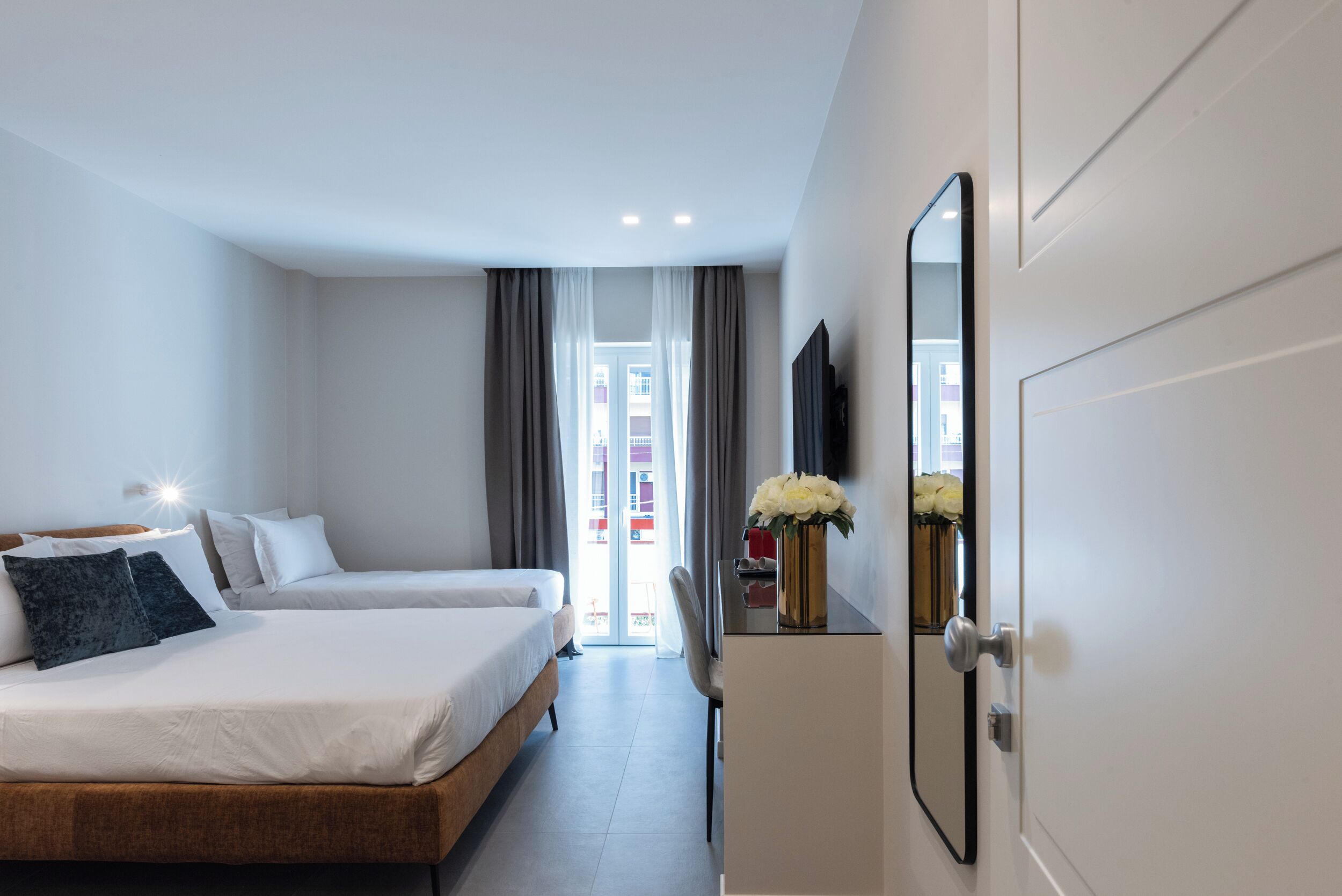 A hotel room with two beds, a desk with a mirror, and a view of the outside through a glass door.