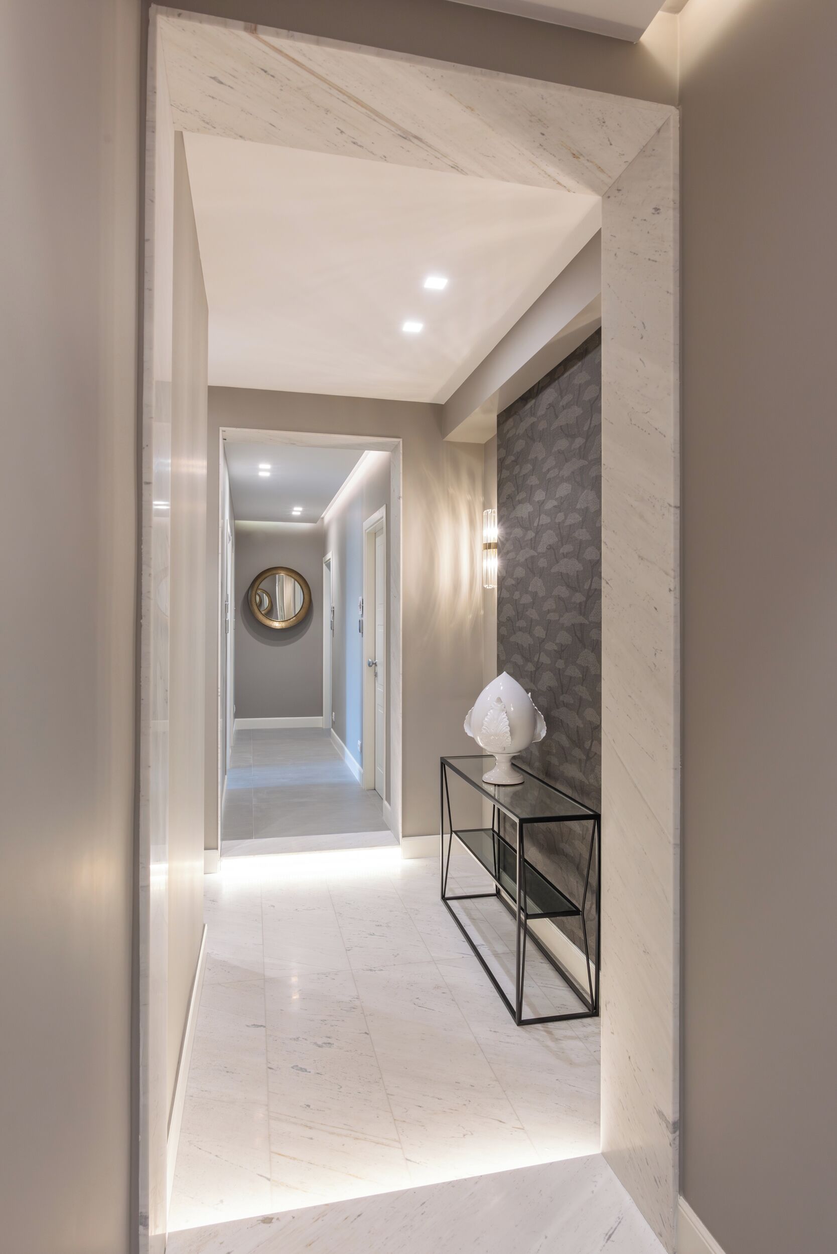 Un couloir moderne avec un sol en marbre, un miroir rond accroché au mur et une petite table sur laquelle est posé un objet décoratif.