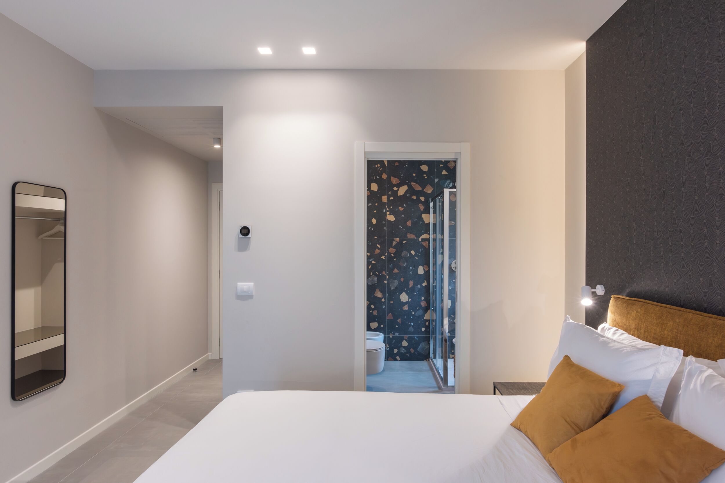 A modern hotel room with a large bed, a mirror, and a door leading to a bathroom with a unique tile backsplash.