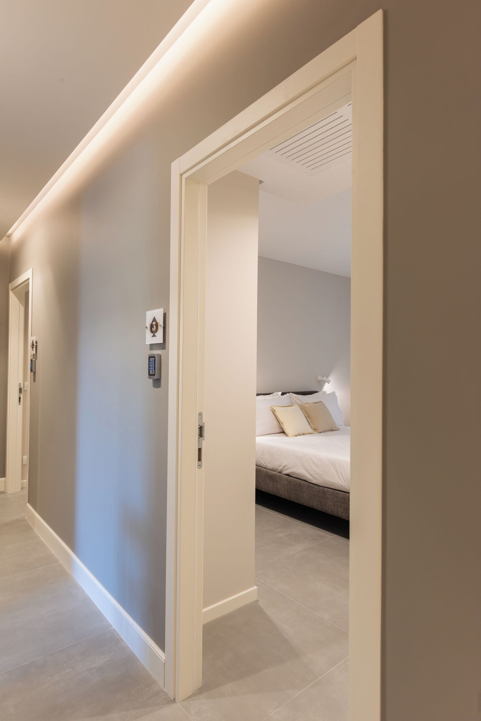 En hotelgang med et moderne design, med en seng med gavl og puder, en dør med kodefelt og en vægmonteret lampe.