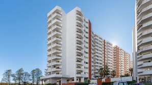 Um edifício residencial moderno e alto, com várias varandas, céu claro e áreas verdes.