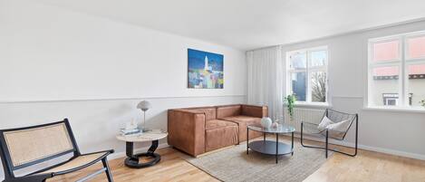 Un salon moderne avec un canapé en cuir marron, un fauteuil noir, une petite table ronde et une grande fenêtre avec des rideaux.