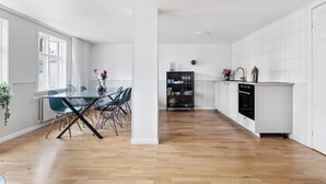 Una cocina moderna con armarios blancos, un comedor con mesa y sillas de vidrio, y vistas de la ciudad a través de amplias ventanas.