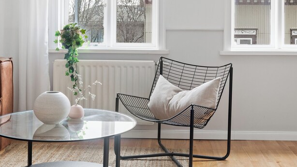 Una habitación con una mesa redonda de vidrio, una silla de marco negro, un radiador blanco y vistas a edificios a través de amplias ventanas.