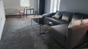 Egy modern nappali, szürke színű kanapéval, üveg asztallappal ellátott kávézóasztallal és egy kisebb asztallal, melyhez székek tartoznak. Két ablak van benne, melyekre redőnyt szereltek, emellett egy radiátor és egy mennyezeti lámpatest is található a helyiségben.