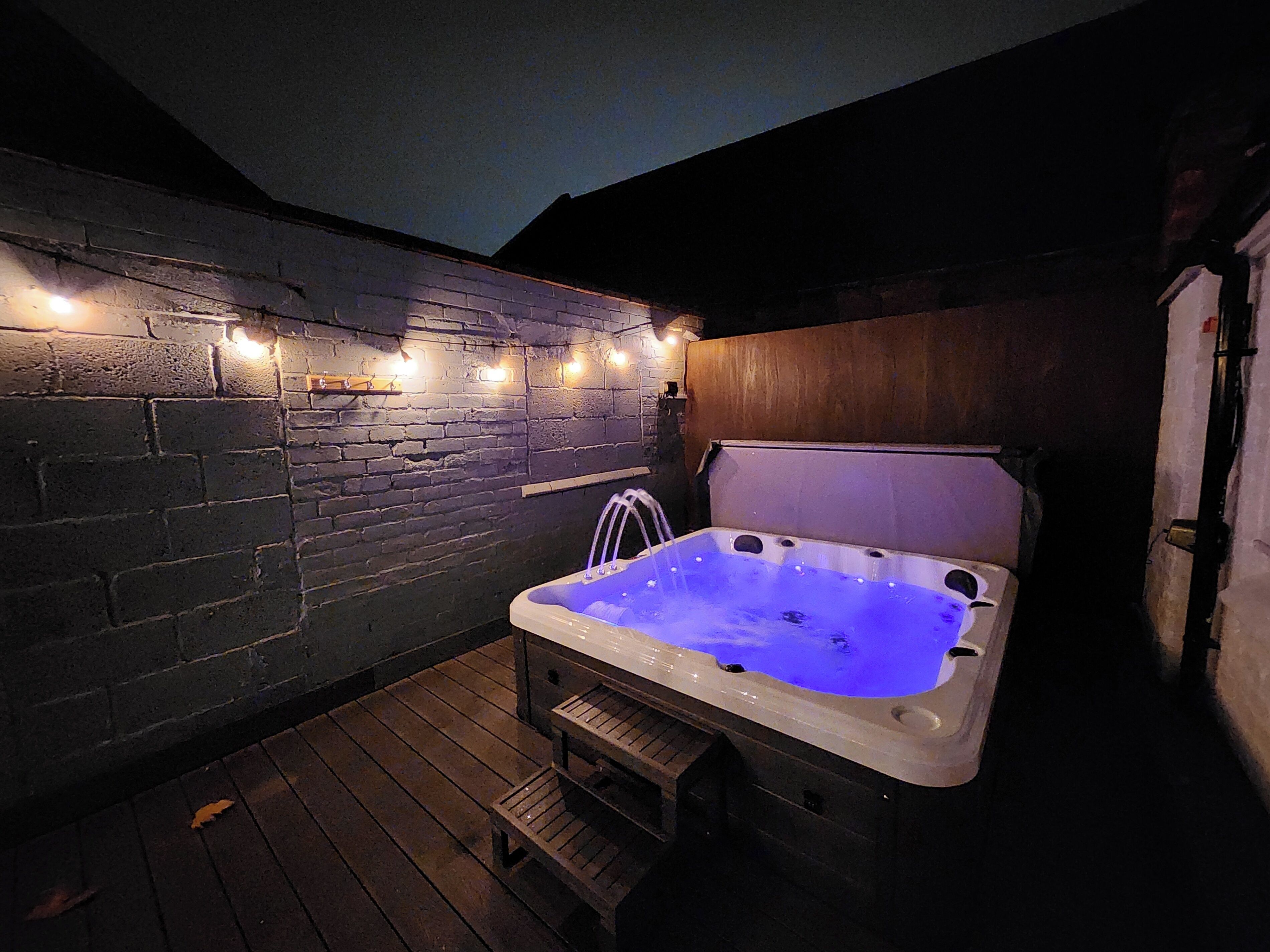 Un bain à remous doté d’une terrasse en bois et de murs en briques, le tout illuminé par des guirlandes lumineuses.