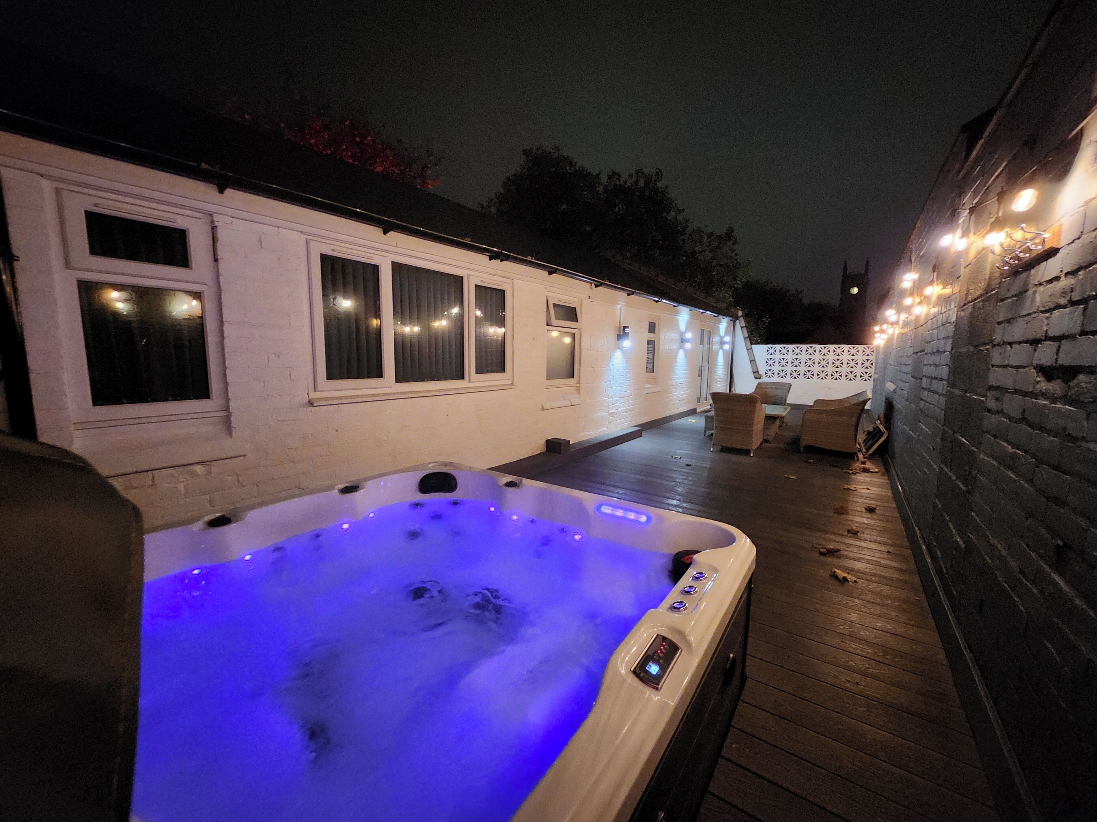 Un bain à remous doté d’un panneau de commande, entouré d’une terrasse en bois et d’un bâtiment blanc avec des fenêtres.