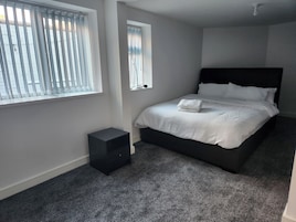 A bedroom with a bed, a small bedside table, a window with blinds, and a carpeted floor.