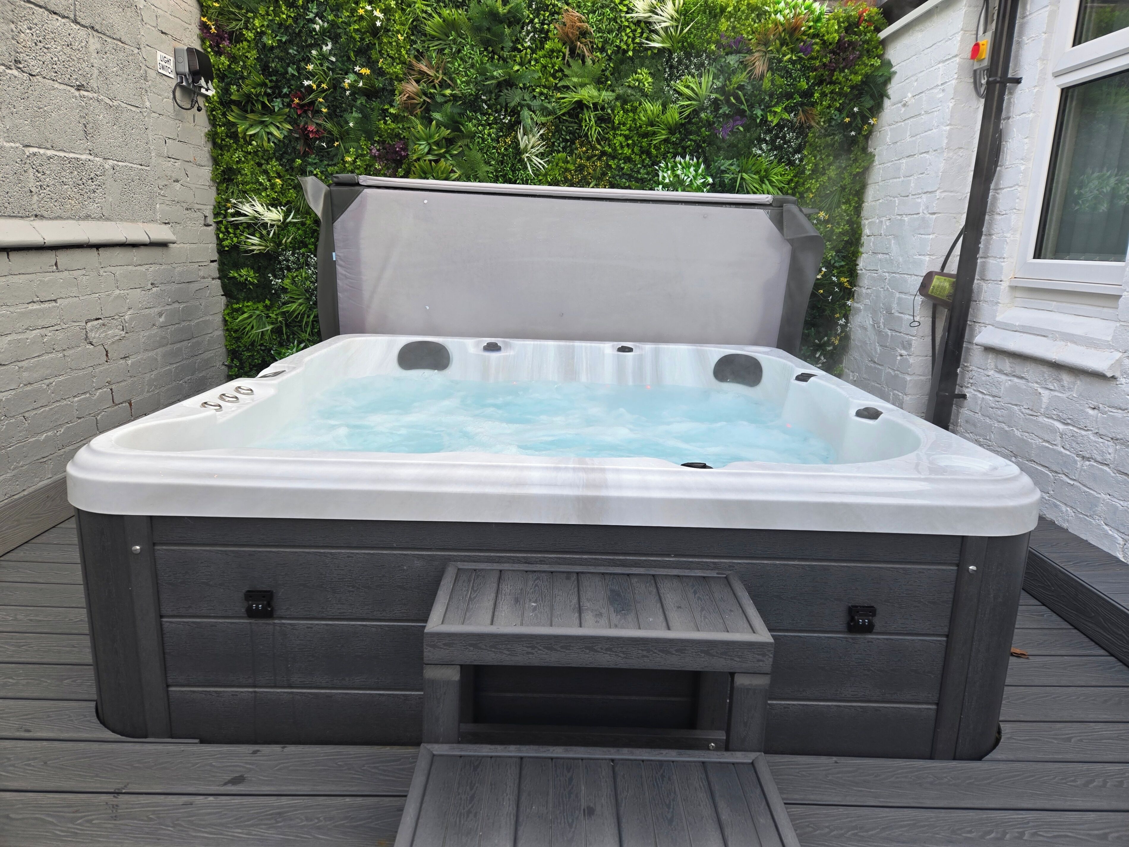 A hot tub with a wooden deck, surrounded by a brick wall and lush greenery.