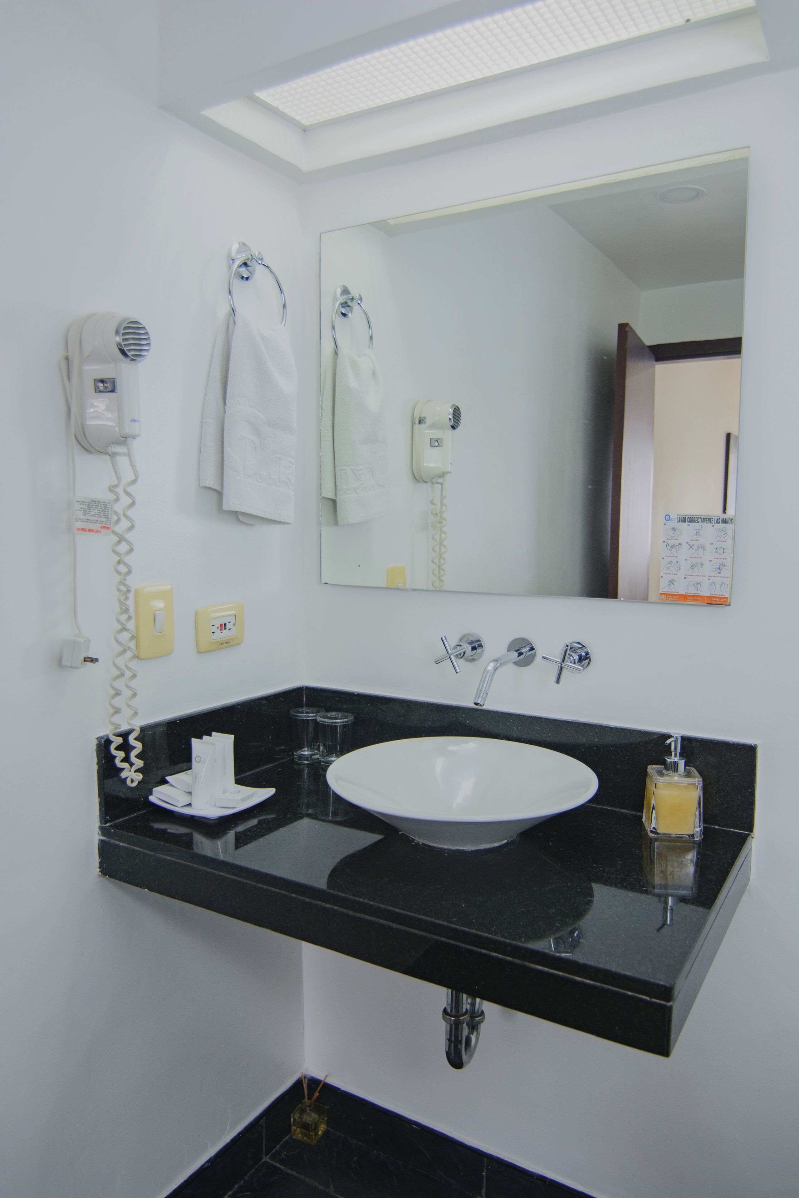 Un baño con encimera negra, lavabo blanco y espejo arriba. Hay dos secadores de pelo en la pared, y se ve un perchero para toallas.