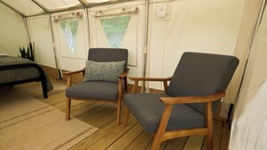 Ein Zelttraum mit zwei Holzstühlen, einem Bett und Blick auf einen Wald durch dreieckige Fenster.