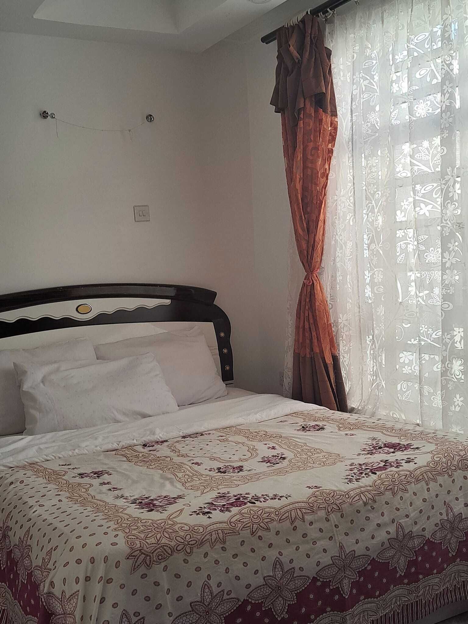 A neatly made bed with a floral-patterned bedspread, a dark headboard, and a window with sheer curtains and a brown curtain.