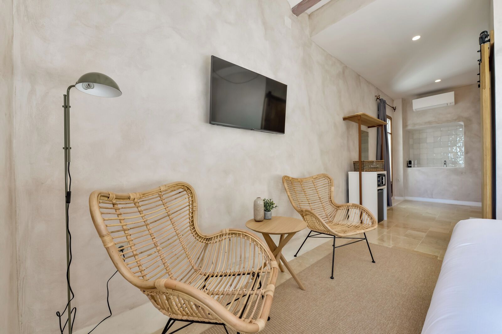 A modern hotel room with a large bed, two wicker chairs, a small round table, a flat-screen TV mounted on the wall, and a wooden shelf.