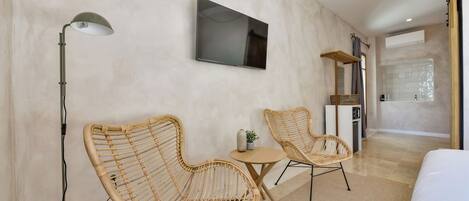 Una habitación de hotel moderna con una cama grande, dos sillones de mimbre, una mesita redonda, un televisor de pantalla plana montado en la pared y un estante de madera.