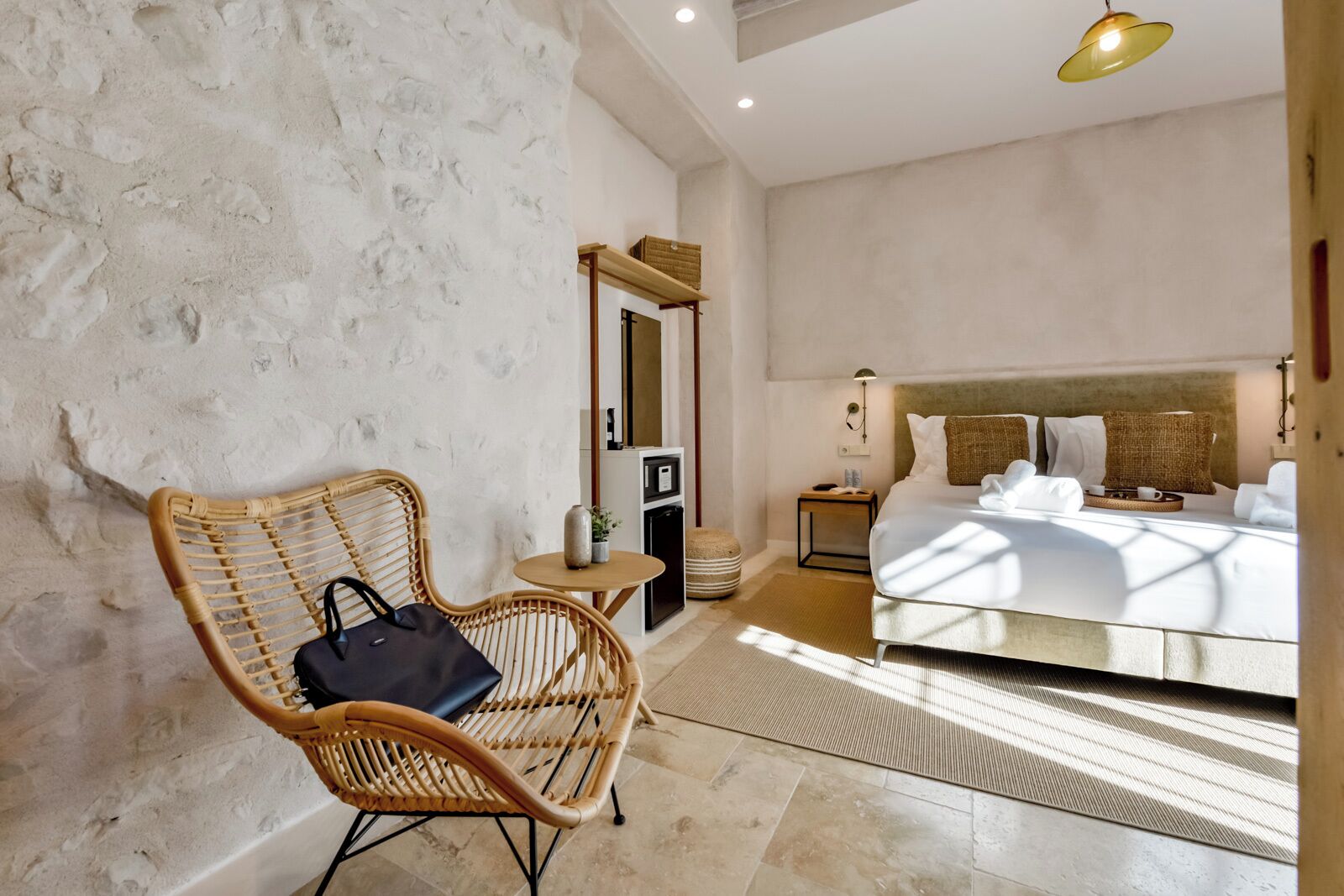 A bedroom with a large bed, a wicker chair, a small table, and a mirror.