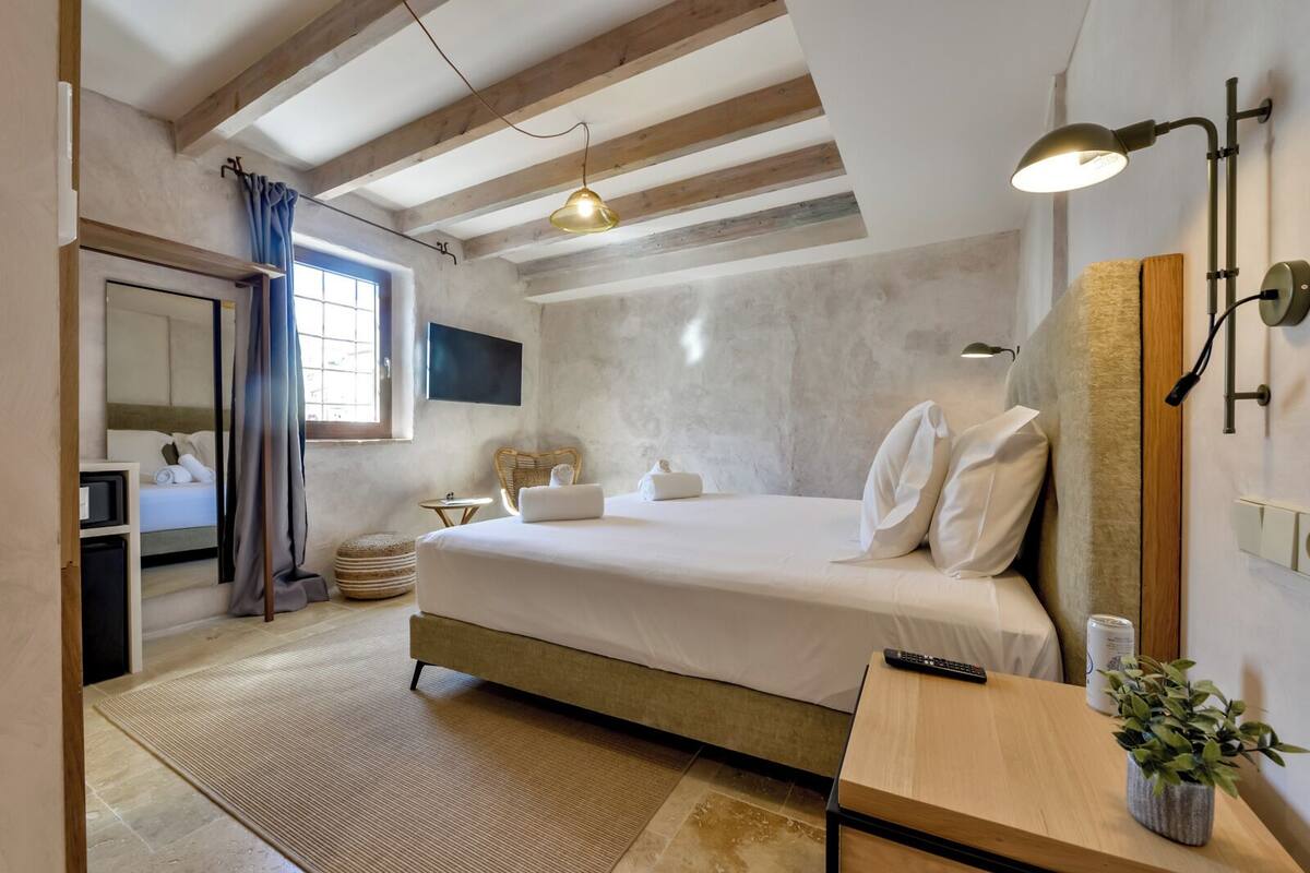Une chambre à coucher moderne avec un grand lit, une table de chevet agrémentée d’une plante, un téléviseur fixé au mur et un luminaire au plafond.