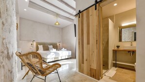 Une chambre moderne avec un grand lit, une porte coulissante en bois donnant sur une salle de bain et un fauteuil en osier.