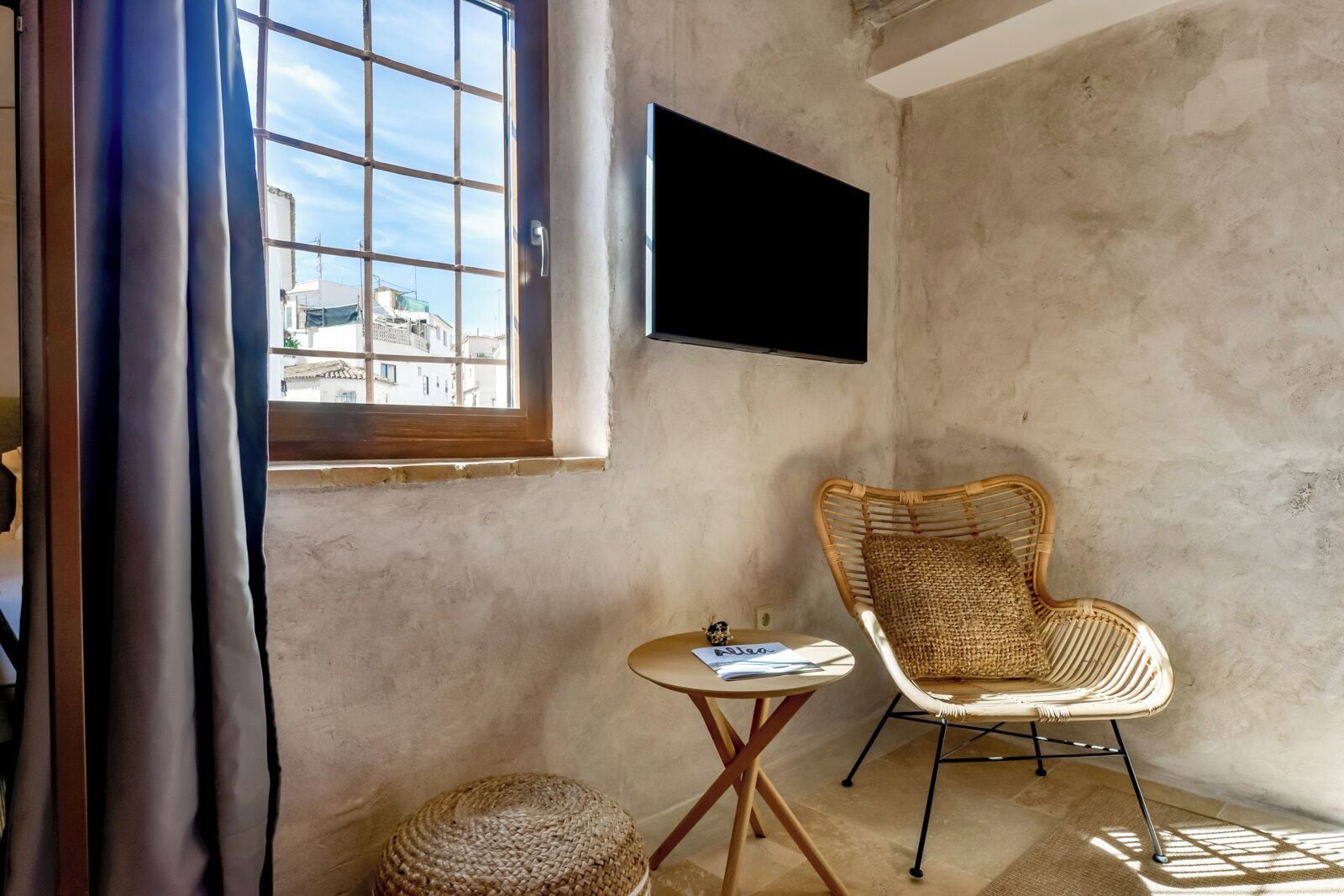 Une pièce dotée d’une grande fenêtre, d’un fauteuil en osier, d’une petite table ronde et d’un téléviseur à écran plat fixé au mur.