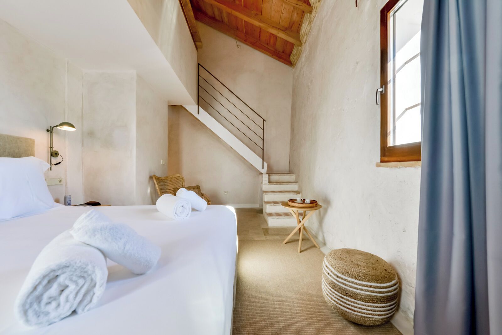Une chambre d’hôtel avec un lit, une petite table, une chaise en osier, un escalier et une fenêtre avec des rideaux.