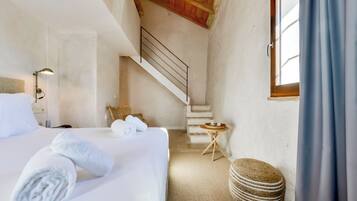 Una camera d'albergo con un letto, un tavolino, una sedia in vimini, una scala e una finestra con tende.