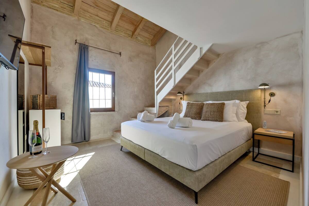 Une chambre à coucher avec un grand lit, une table en bois, une petite table, une table de chevet, une fenêtre avec des rideaux et un escalier.