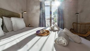 Une chambre d’hôtel avec un lit, une chaise en osier, un plateau avec une tasse et une assiette, et une fenêtre avec des rideaux.