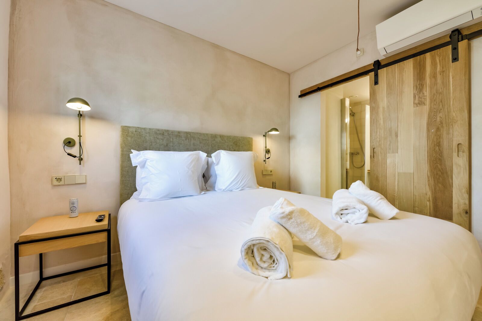 Habitación de hotel con una cama grande, dos mesitas de noche, una lámpara de pared y una puerta corredera hacia la zona de ducha.