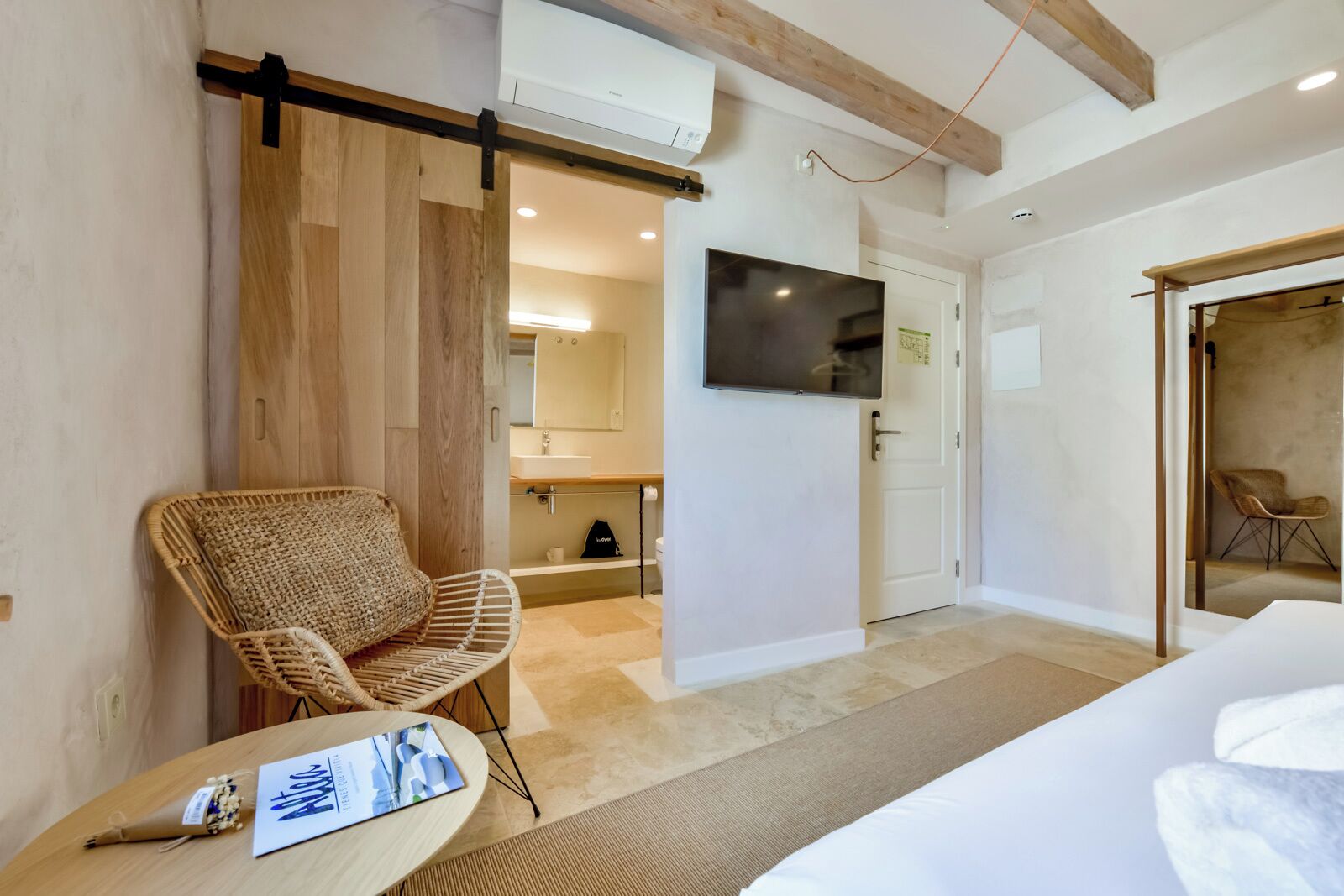 A hotel room with a bed, a wicker chair, a TV, and a bathroom visible through an open door.