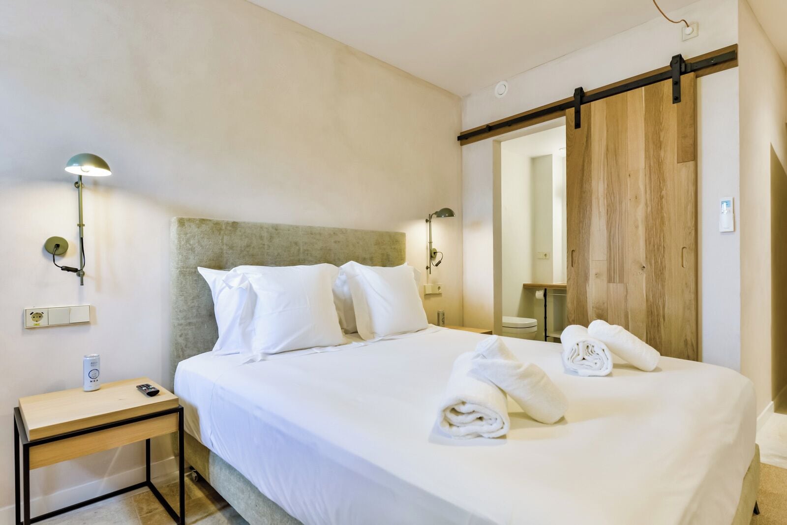 Una cama bien hecha con sábanas blancas, una mesita de noche con lámpara y un armario de madera con puerta corredera.