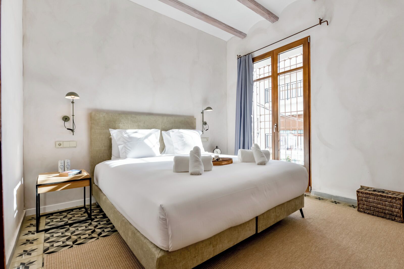 Quarto de hotel com uma cama grande, uma mesa de cabeceira com abajur, uma mesinha com livros e uma janela com vista para edifícios.
