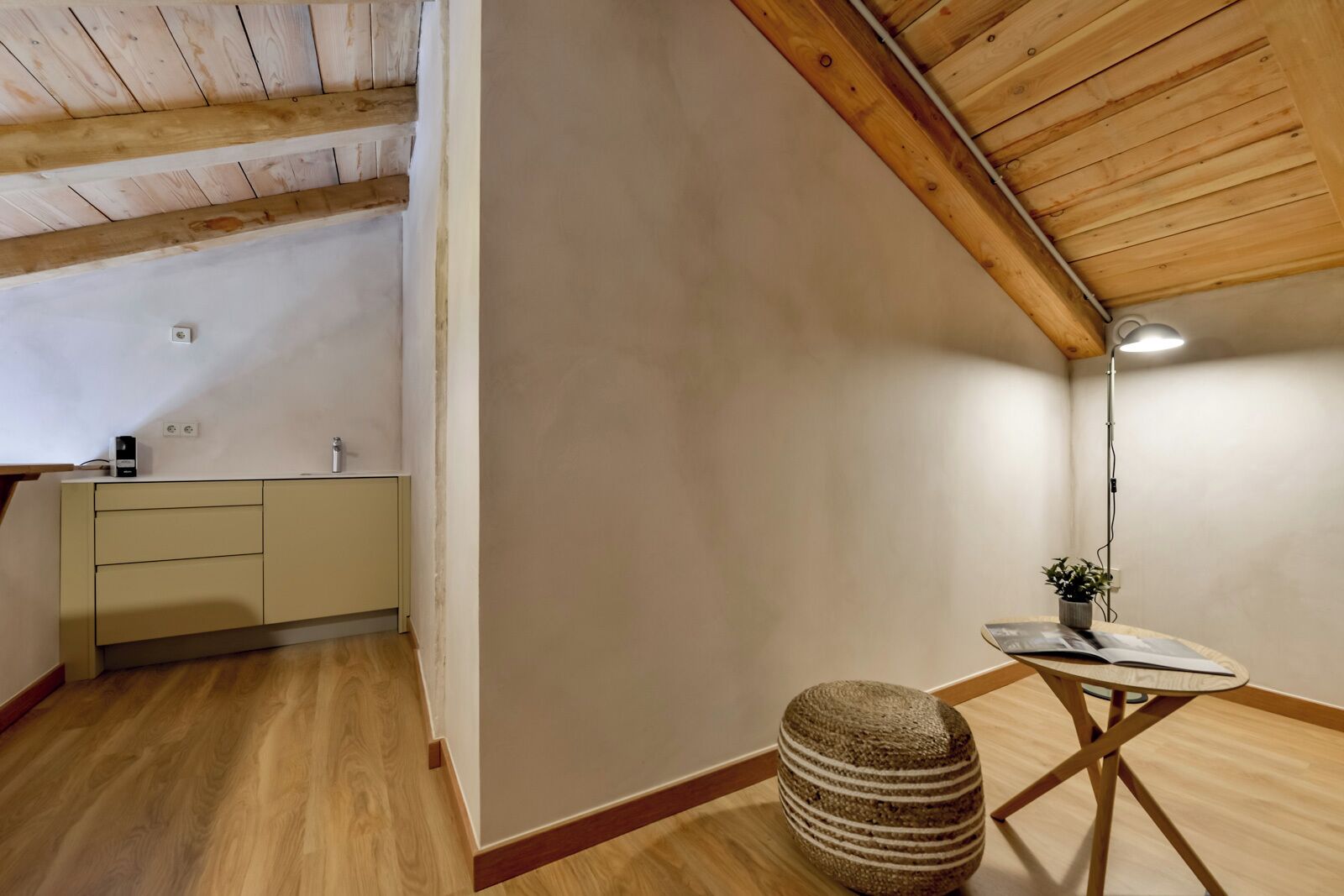 A small, cozy room with a wooden ceiling, a kitchenette with a sink and cabinets, a small round table with a plant, and a woven stool.