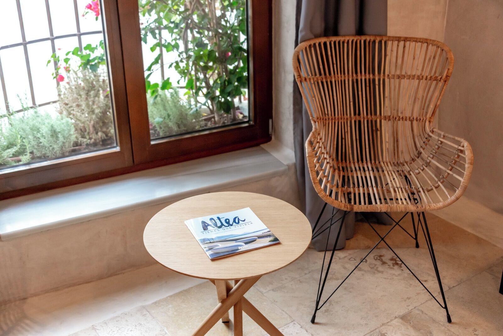 A room with a window, a wicker chair, a small round table with a magazine, and a view of greenery outside.