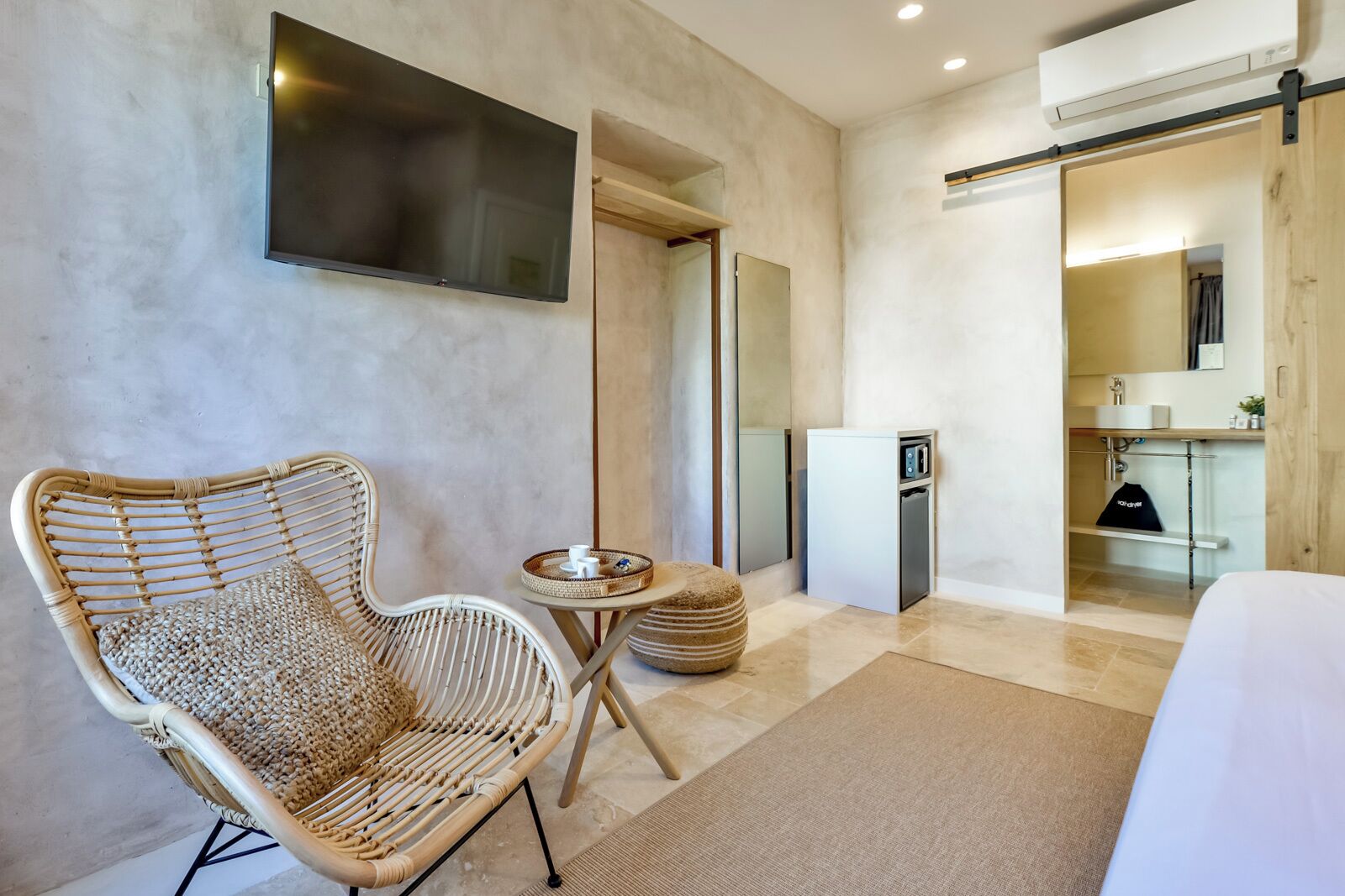 A modern hotel room with a large flat-screen TV, a wicker chair, a small round table, and a compact kitchenette.