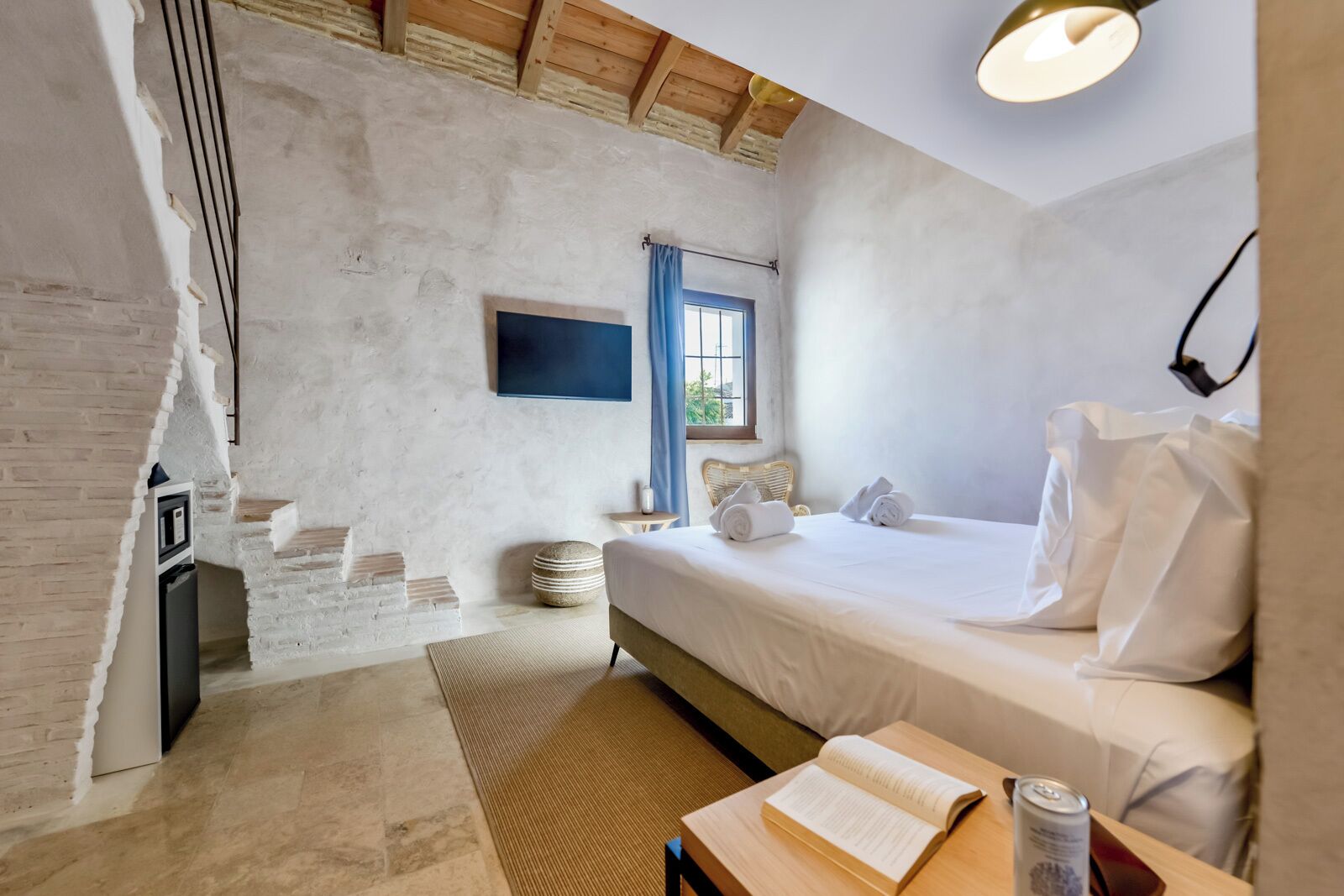 A bedroom with a bed, a bedside table with a book and a can, a window with blue curtains, a wall-mounted TV, and a brick wall.
