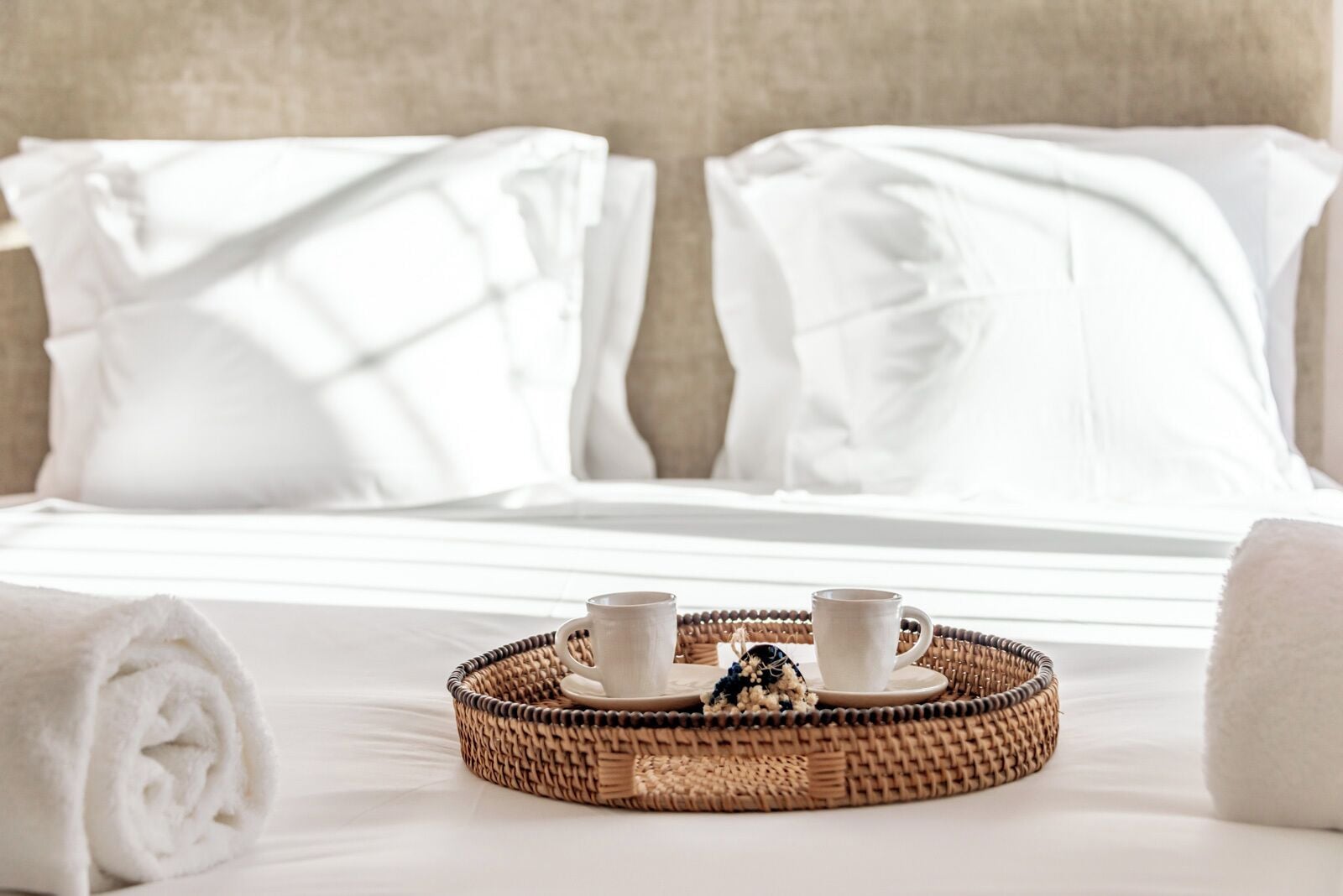 A neatly made bed with white linens, a tray with two cups and a small decorative item, and rolled towels on the bed.