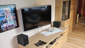 Uma sala de estar moderna com um painel de entretenimento em madeira, uma televisão de tela plana e diversos objetos decorativos, incluindo plantas e um quadro emoldurado.
