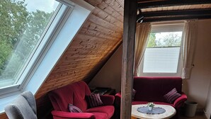Um quarto em estilo loft com teto inclinado, piso de madeira, uma poltrona cinza, um sofá vermelho e uma pequena mesa redonda com uma planta.